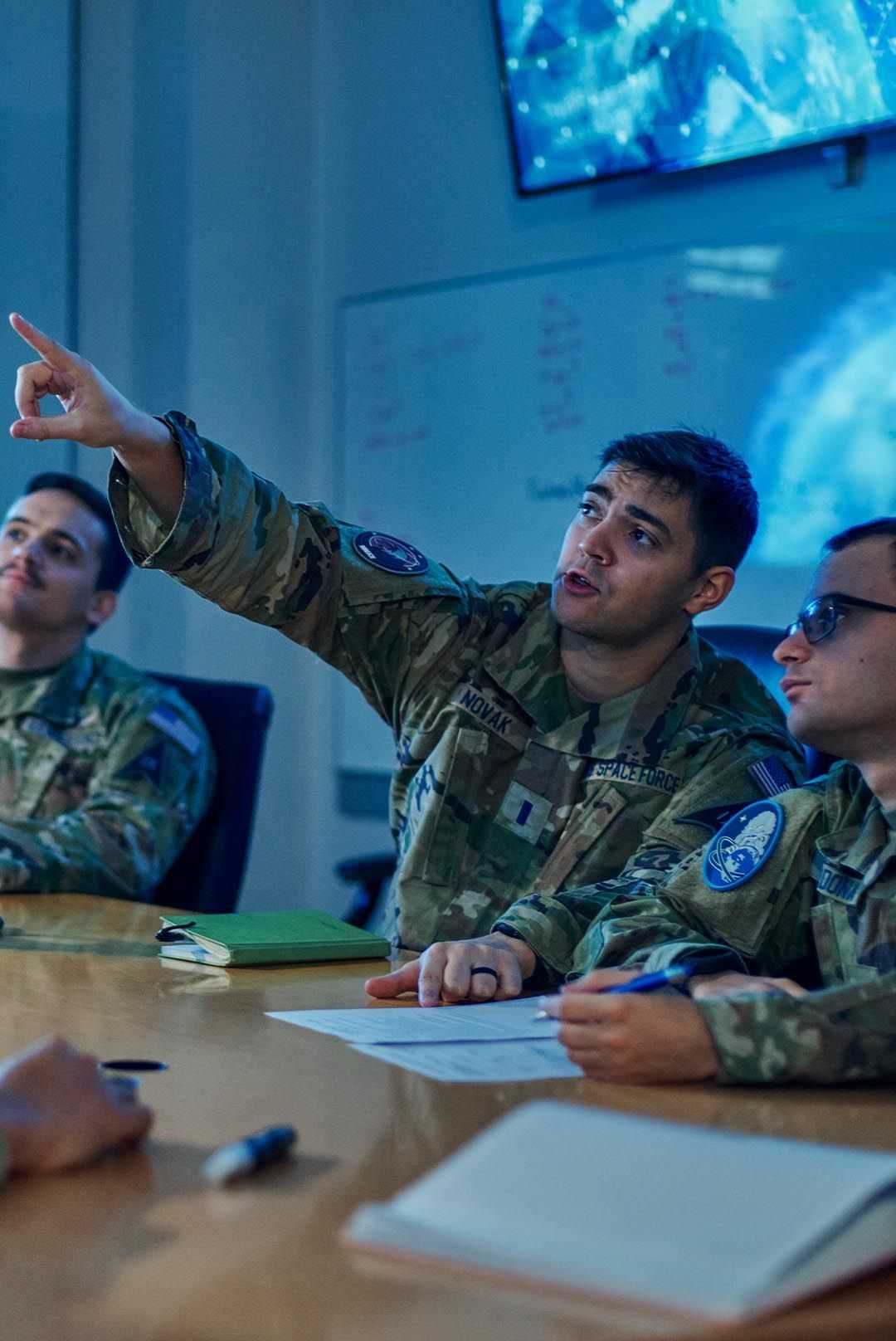 Uniformed military members gathered around a table, engaged with a screen in front of them