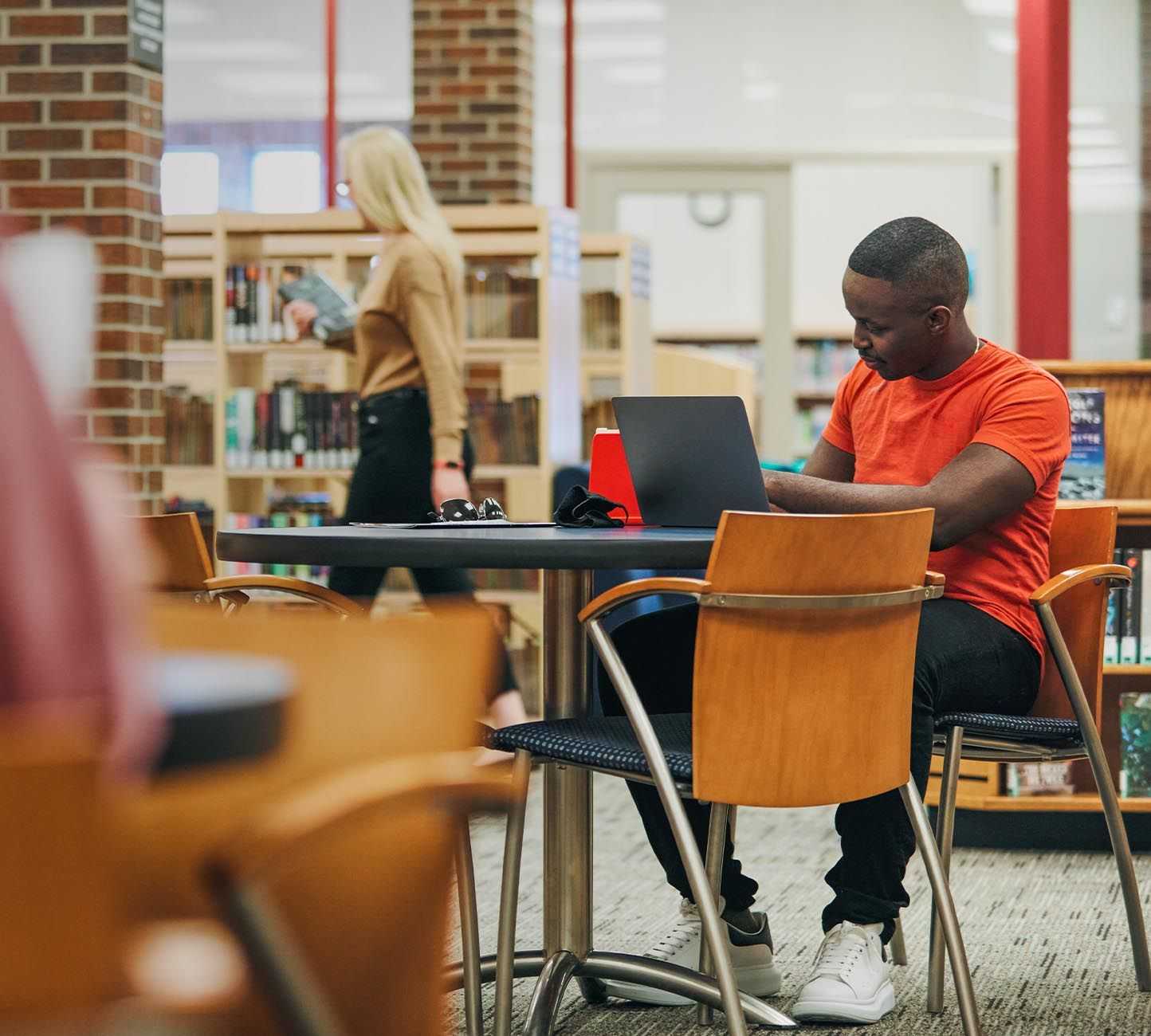A man seated at a library table, focused on his laptop, surrounded by books